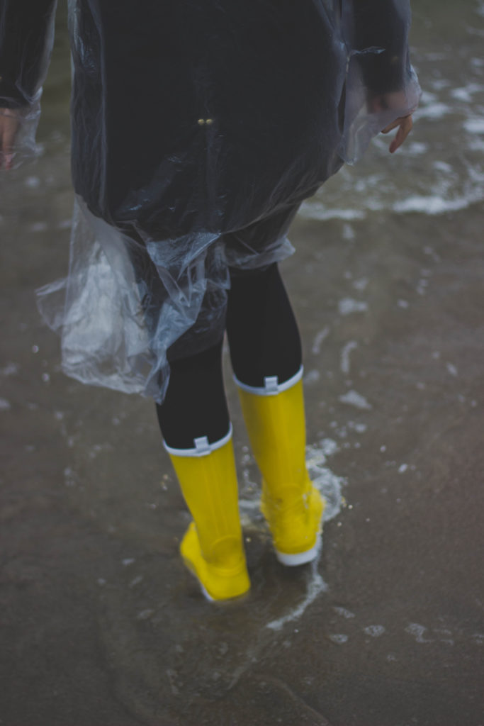 Woman in rain gear shown from the waist down, trudging through water in yellow rubber boots. Asheville water damage is a strong possibility.